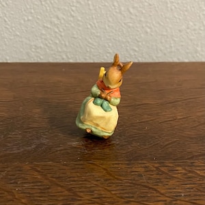 May include: A small ceramic figurine of a brown rabbit wearing a green dress and sitting in a yellow chair. The rabbit is holding a green ball of yarn in its paws.