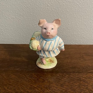 May include: A ceramic figurine of a pig wearing blue and white striped pajamas and carrying a green and yellow cabbage. The pig has a pink face and a small, pink snout.