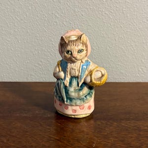 May include: A ceramic figurine of a cat dressed in a bonnet and apron, holding a basket. The cat has green eyes and is wearing a pink bonnet, a blue and white apron, and a skirt with pink polka dots. The figurine is on a wooden surface.