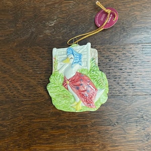 Può includere: Un ornamento in ceramica con Jemima Puddle-Duck dal libro per bambini di Beatrix Potter. L'ornamento raffigura Jemima con un vestito rosso e bianco, un berretto blu e scarpe gialle, in piedi in un giardino verde. L'ornamento è attaccato a un cordoncino dorato.