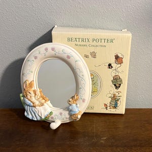 May include: Oval mirror with a cream frame, decorated with flowers and bunny rabbit figures. The mirror is from the Beatrix Potter Nursery Collection. A box with Beatrix Potter character illustrations sits beside the mirror.