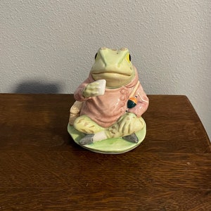 Puede incluir: Una figurita de cerámica de una rana que lleva una túnica rosa y pantalones verdes, sosteniendo un objeto blanco en su mano. La rana está sentada sobre una base verde.