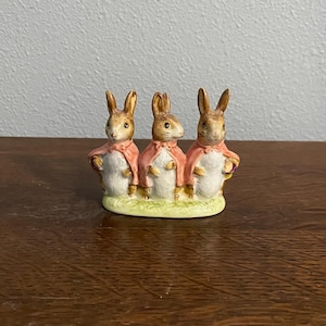 May include: A ceramic figurine featuring three rabbits wearing pink capes. The rabbits have brown ears and faces, and are standing on a green base. The figurine is likely a decorative item or collectible.