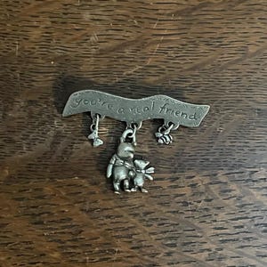 May include: A silver-toned pin featuring the phrase "You're a real friend." Below the inscription are dangling charms of a flower, Winnie the Pooh and Piglet, and a bee. The pin is likely a collectible or a piece of jewelry.