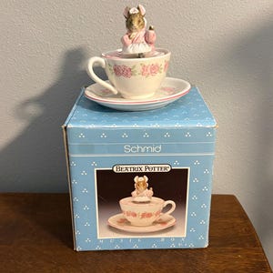 Puede incluir: Una caja de música Schmid Beatrix Potter con una taza y un platillo con diseño floral. Una figura de un ratón con un vestido rosa se encuentra encima de la taza. La caja azul tiene lunares blancos y las palabras "Schmid" y "Beatrix Potter".