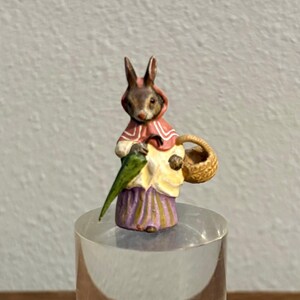 May include: A miniature figurine of a rabbit dressed in a red hooded cloak and a yellow and purple dress, holding a green umbrella and a woven basket. The rabbit has brown fur and is standing on a clear, cylindrical base.