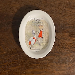 Puede incluir: Un plato de cerámica blanco con una imagen de Jemima Puddle-Duck del libro de Beatrix Potter "The Tale of Jemima Puddle-Duck". El plato incluye una cita del libro: "BUT as to a nest-there is no difficulty."