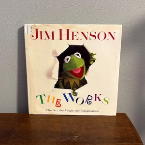 May include: A book titled "Jim Henson The Works" with Kermit the Frog peeking through a torn hole in the cover. The book's title is in red, and the subtitle "The Art, the Magic, the Imagination" is below the title in a colorful font.
