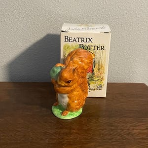 May include: A small ceramic figurine of a squirrel holding a green acorn. The figurine is brown and white with a green base. The figurine is in a box with the text "Beatrix Potter" on it.