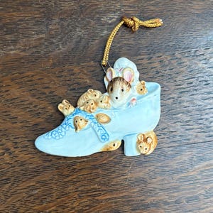 May include: A whimsical ornament featuring a light blue shoe filled with several small mice. The mice are tan and white, with some wearing blue blankets. A gold cord is attached for hanging. The ornament is likely ceramic.