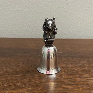 May include: A silver-colored bell with a detailed figurine of a mouse wearing a dress on top. The bell has a polished, reflective surface and sits on a wooden surface. The mouse figurine is intricately detailed.