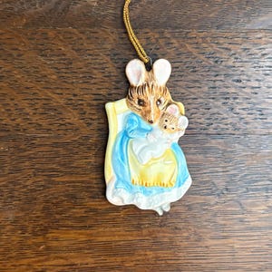 Può includere: Un ornamento in ceramica raffigurante una mamma topo che tiene in braccio un topolino. La mamma topo indossa un vestito blu e giallo. L'ornamento ha un cordino dorato per appenderlo. Lo sfondo è una superficie di legno.