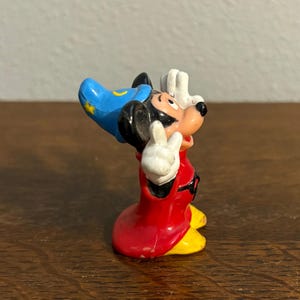 May include: A small figurine of Mickey Mouse in a sorcerer's outfit. The figure has a red robe, yellow shoes, a blue hat with a yellow crescent moon and star, and white gloves. Mickey is posed with his hands raised in a gesture.