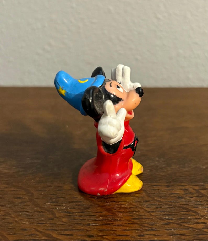 May include: A small figurine of Mickey Mouse in a sorcerer's outfit. The figure has a red robe, yellow shoes, a blue hat with a yellow crescent moon and star, and white gloves. Mickey is posed with his hands raised in a gesture.