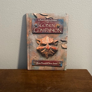 Puede incluir: Libro « A Field Guide to Goblins: The Goblin Companion ». La cubierta texturizada presenta una cara de duende y texto. El libro tiene un aspecto rústico y envejecido con tonos azules, marrones y rojos. Por Brian Froud & Terry Jones.