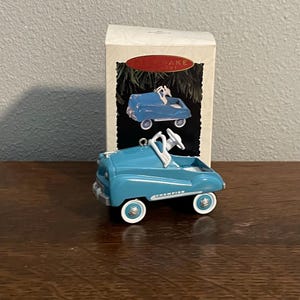 May include: A blue and white vintage toy car ornament with the word "Champion" on the side. The ornament is in its original box, which is white with red trim and the word "Keepsake" on it.