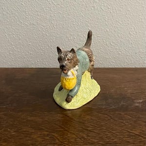 Puede incluir: Figura de cerámica de un perro con una chaqueta azul y una cesta amarilla. El perro es marrón y blanco, de pie sobre una base verde. La figura está sobre una superficie de madera.