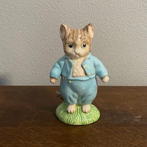 Puede incluir: Una figura de cerámica de un gato con un atuendo azul claro con botones amarillos. El gato está de pie sobre una base verde y cubierta de hierba. El gato tiene pelaje marrón y beige.