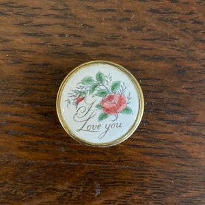 Puede incluir: Una caja redonda de cerámica blanca con un borde dorado. La tapa presenta una rosa rosa con hojas verdes y las palabras "Te amo" en cursiva.