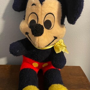 May include: Vintage Mickey Mouse plush toy. The plush features a black body, ears, and arms, with a yellow face, hands, and feet. Mickey wears red shorts with yellow buttons and a yellow bow tie. The toy is a classic Disney collectible.