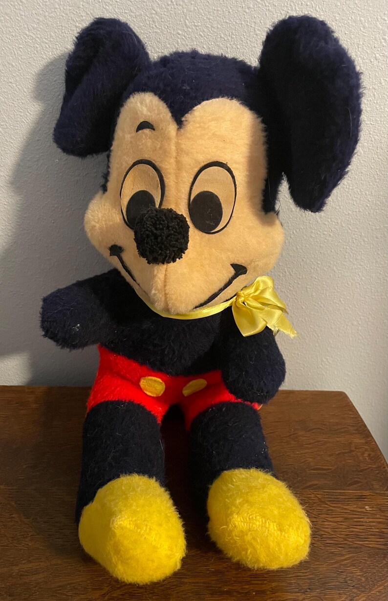 May include: Vintage Mickey Mouse plush toy. The plush features a black body, ears, and arms, with a yellow face, hands, and feet. Mickey wears red shorts with yellow buttons and a yellow bow tie. The toy is a classic Disney collectible.