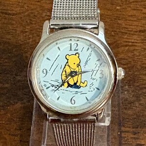 May include: A silver wristwatch with a light blue face featuring a yellow Winnie the Pooh character. The watch has a silver mesh band and black hands. The watch face has black numbers and markings.