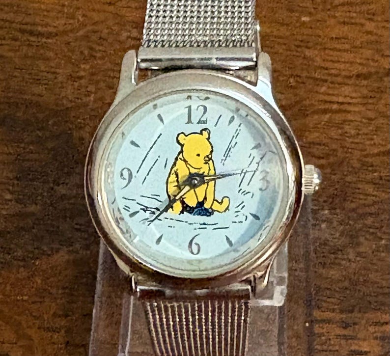 May include: A silver wristwatch with a light blue face featuring a yellow Winnie the Pooh character. The watch has a silver mesh band and black hands. The watch face has black numbers and markings.