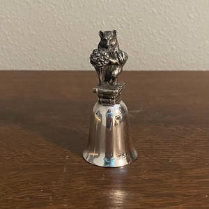 May include: A silver bell with a squirrel figurine on top. The squirrel is holding a bunch of flowers and is standing on a small base.