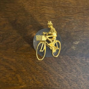 Op de afbeelding: Een goudkleurige fietspen met een fietser met een mand. De fietser draagt een helm en rijdt op een fiets met een mand aan de voorkant. De pin is geplaatst op een heldere, ronde basis.
