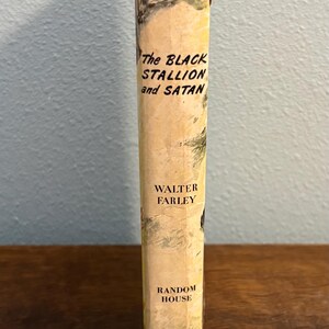 May include: A vintage book titled "The Black Stallion and Satan" by Walter Farley. The book's spine is a light beige with black text and a black top. The publisher is Random House.