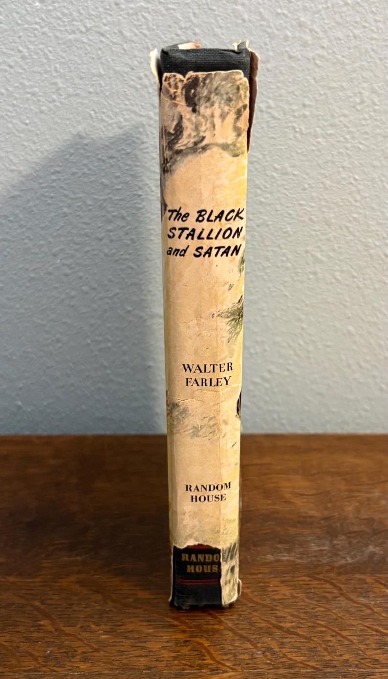 May include: A vintage book titled "The Black Stallion and Satan" by Walter Farley. The book's spine is a light beige with black text and a black top. The publisher is Random House.