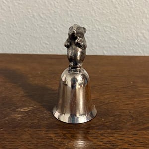 May include: A silver-colored bell with a detailed figurine of a small animal on top. The bell has a flared base and a smooth, reflective surface. The animal figure is in a standing position, adding a whimsical touch to the design.