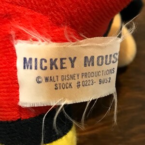 May include: A white tag with the words "MICKEY MOUSE" in blue block letters. The tag also includes "&copy; WALT DISNEY PRODUCTIONS" and "STOCK #0223- 9052". The tag is attached to a red plush toy.