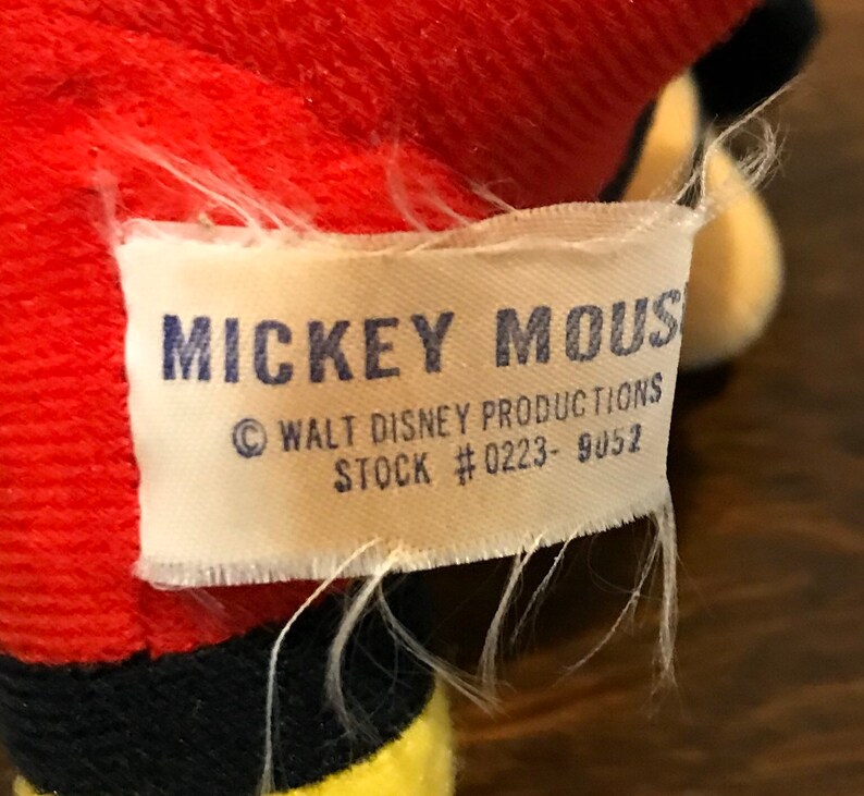 May include: A white tag with the words "MICKEY MOUSE" in blue block letters. The tag also includes "&copy; WALT DISNEY PRODUCTIONS" and "STOCK #0223- 9052". The tag is attached to a red plush toy.