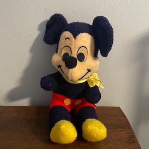 May include: A vintage Mickey Mouse plush toy. The plush features a black head and ears, a yellow face, and a black nose. It wears red shorts with yellow buttons, yellow feet, and a yellow bow tie. The toy is sitting on a wooden surface.