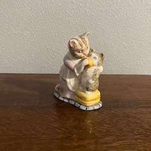 Puede incluir: Figura de cerámica que representa dos gatos. Uno, con un vestido rosa, abraza a otro. El segundo gato sostiene un objeto amarillo. La figura está sobre una base con un borde azul y gris.