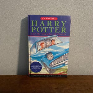 May include: A hardcover book titled "Harry Potter and the Chamber of Secrets" by J.K. Rowling. The cover features a blue car with Harry, Ron, and a pet owl. The book is on a wooden surface.