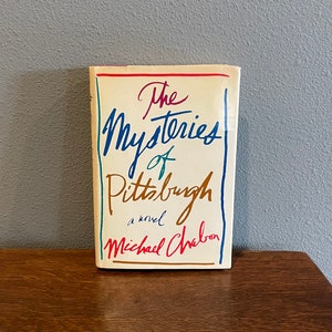 May include: A book titled "The Mysteries of Pittsburgh" by Michael Chabon. The book cover is white with colorful handwritten text.