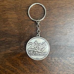 Pode incluir: Chaveiro de prata com um charme redondo de metal com Mickey Mouse e seus amigos. O charme diz "20th Walt Disney World. Celebrate the Future Hand in Hand."