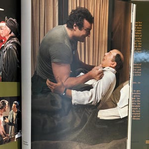 Op de afbeelding: Een sc&egrave;ne uit een film of toneelstuk. Een man in een grijs t-shirt houdt een andere man in een wit overhemd vast. De sc&egrave;ne speelt zich af in een kamer met een bed en gordijnen, wat wijst op een interieur.