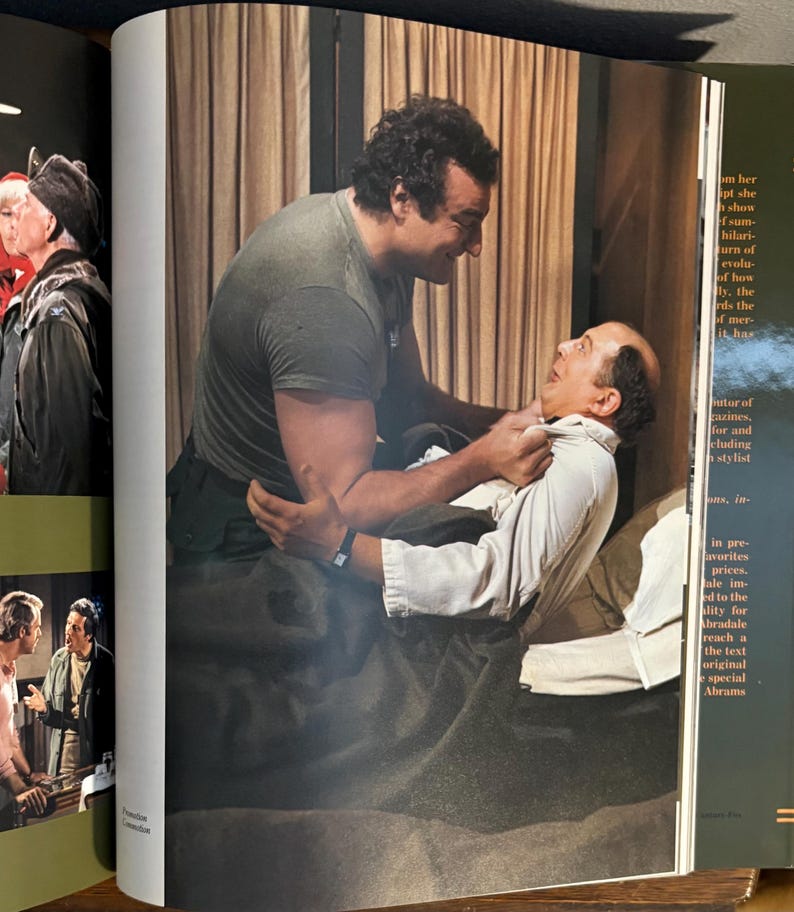 Op de afbeelding: Een sc&egrave;ne uit een film of toneelstuk. Een man in een grijs t-shirt houdt een andere man in een wit overhemd vast. De sc&egrave;ne speelt zich af in een kamer met een bed en gordijnen, wat wijst op een interieur.