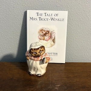 May include: A ceramic figurine of Mrs. Tiggy-Winkle wearing a white bonnet and a red and white checkered blouse, positioned in front of a white book titled The Tale of Mrs. Tiggy-Winkle by Beatrix Potter with a matching hedgehog illustration.