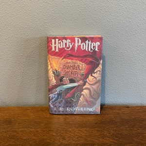 May include: A red, white, and black paperback book titled "Harry Potter and the Chamber of Secrets" by J.K. Rowling. The cover features an illustration of Harry Potter fighting a snake.