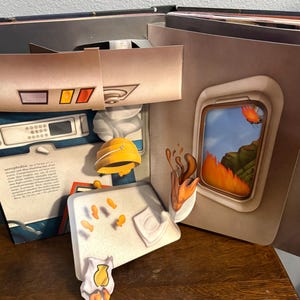May include: A pop-up book with a theme of aerophobia. The open pages reveal a plane interior, a window with a fiery landscape, and other related illustrations. The book features a yellow hard hat and other paper cutouts.
