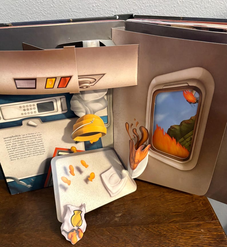 May include: A pop-up book with a theme of aerophobia. The open pages reveal a plane interior, a window with a fiery landscape, and other related illustrations. The book features a yellow hard hat and other paper cutouts.