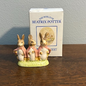 May include: A ceramic figurine featuring three rabbits wearing pink capes, standing on a patch of green grass. The figurine is from "The World of Beatrix Potter" collection, with the box visible in the background.