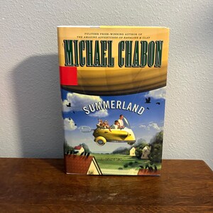 May include: A hardcover book titled "Summerland" by Michael Chabon. The cover features a yellow car with passengers flying over a landscape with houses and trees. The author's name is in large, bold letters.