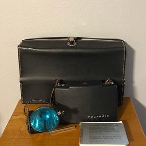 Może przedstawiać: Winietkowy aparat Polaroid z czarnym skórzanym futerałem, niebieską lampą błyskową i instrukcją obsługi zatytułowaną "Polaroid Cold-Clip 403".