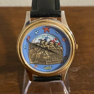 May include: A gold-toned wristwatch with a black leather band. The watch face features a light blue background with a bronze-colored relief of people on a boat, surrounded by sea creatures. The watch has a gold-colored bezel.