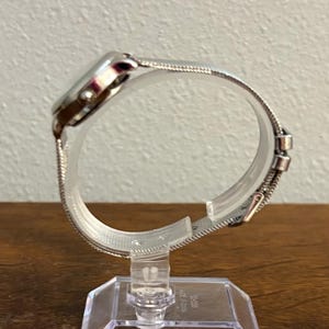 May include: A silver-toned wristwatch with a mesh band. The watch face is round and the band is secured with a clasp. The watch is displayed on a clear plastic stand.